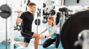 A female personal trainer assisting a young male lifting a dumbbell while bending his knees